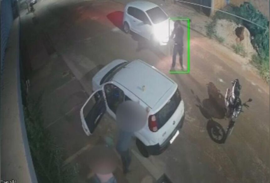 Os três descem do carro quando são surpreendidos por um homem em uma motocicleta, que retira uma arma da cintura e começa a disparar contra a vítima